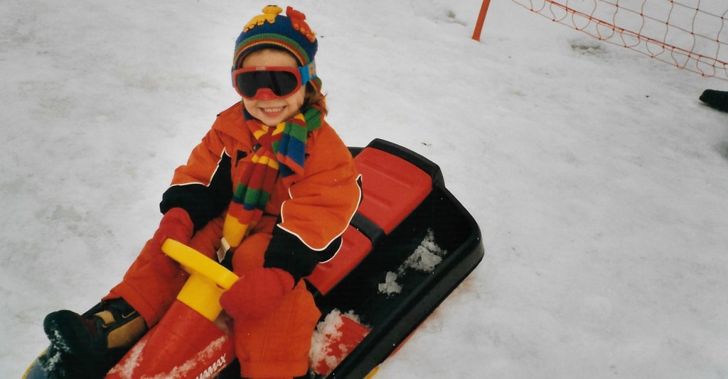 A child sits on a sled in a colorful ski suit and looks cheekily into the camera.
